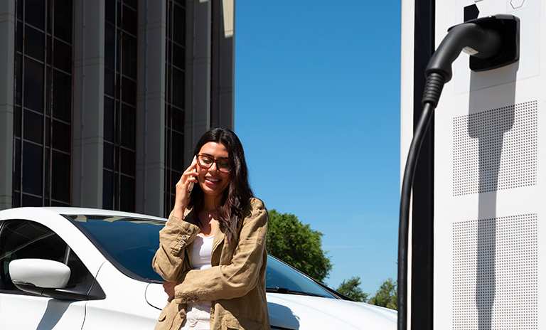 Mujer hablando por celular mientras carga su vehículo eléctrico. 