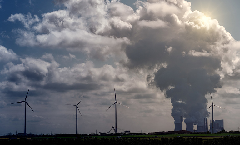 Empresa de la que sale humo que se dirige a la amósfera. Al lado hay molinos de viento.