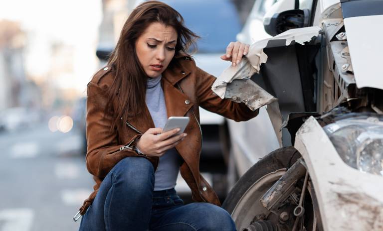 Carro accidentado y una mujer recostada en él, triste y mirando su celular.